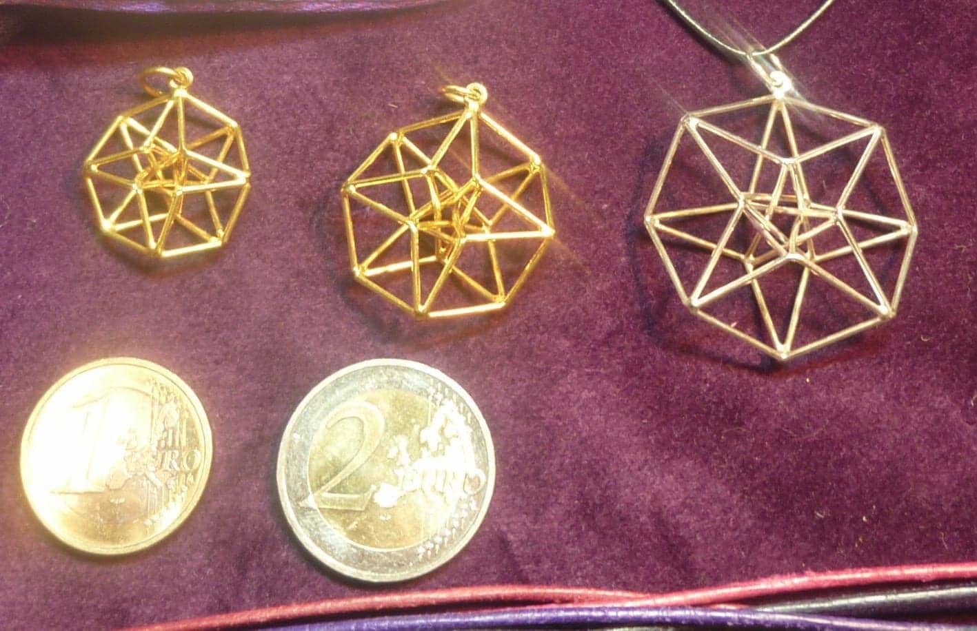 May include: Three geometric pendant necklaces, two gold and one silver, with intricate wireframe designs. Two euro coins are also visible, providing a size reference. The pendants are displayed on a purple surface.